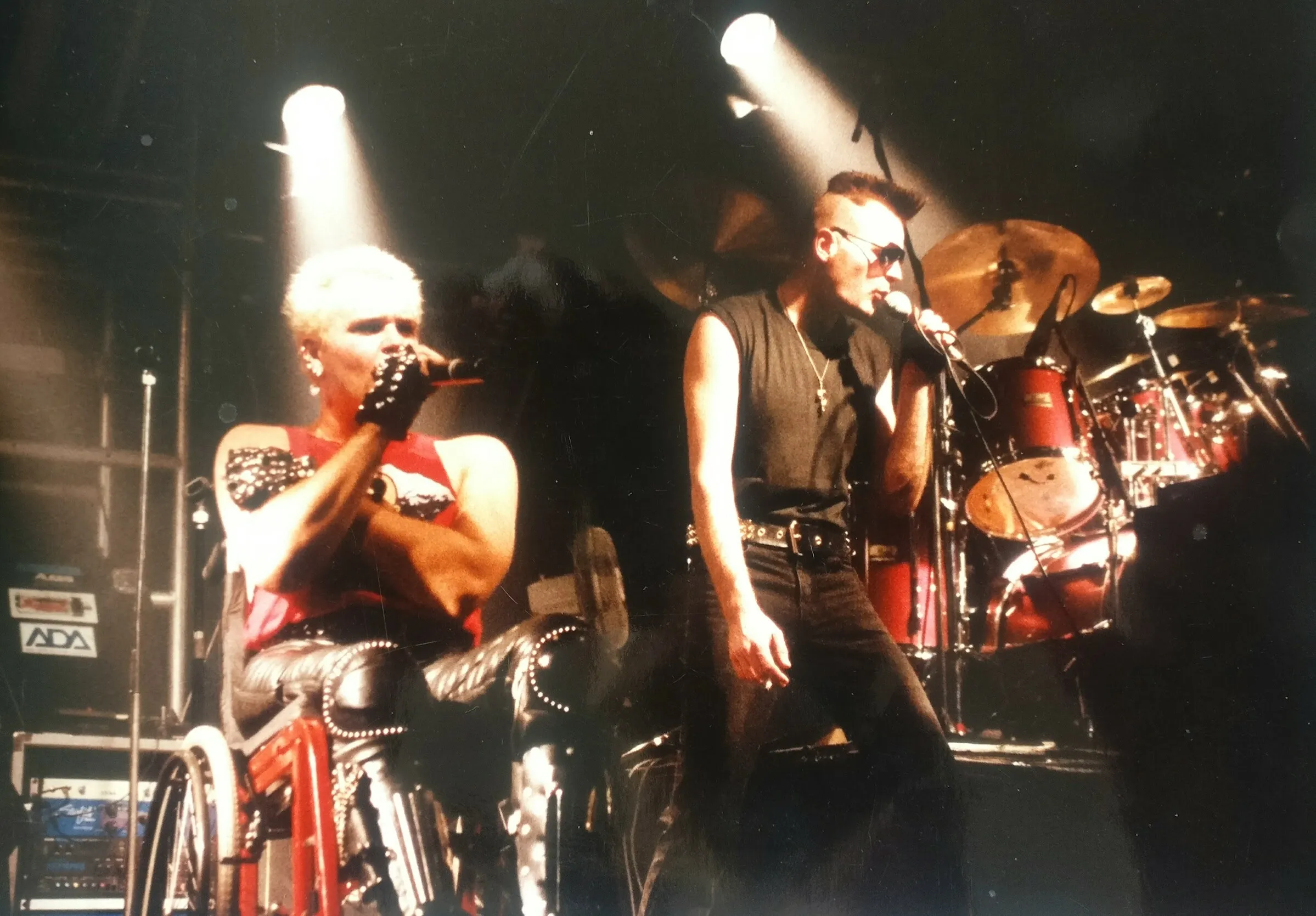 Fotograf&iacute;a. Concierto en directo del grupo Freak UK. En primer plano, dos vocalistas bajo focos cenitales. A tu izquierda, Mik Scarlet canta desde su silla de ruedas, con est&eacute;tica punk: ropa de cuero, guantes y pelo corto decolorado. A la derecha, Tony canta de pie, con vestimenta negra &mdash;vaqueros y camiseta negra&mdash; y el pelo rapado con cresta. Al fondo se distingue la bater&iacute;a.